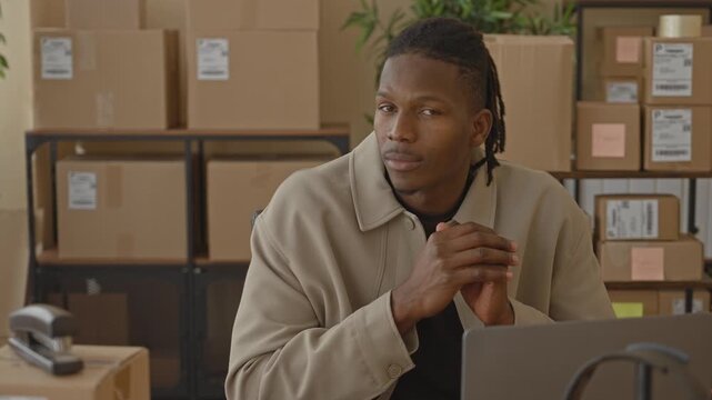 Man with packages clasping hands over laptop in a storage building with stacked boxes and scanner visible; small business pride calm.
