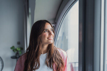 Fototapeta premium Smiling young woman gazes out a large window in a modern apartment, bathed in soft natural light. The casual, reflective portrait conveys optimism, relaxation and quiet morning thoughts.