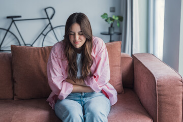 Obraz premium Young woman sitting on couch holding her stomach in pain and discomfort at home