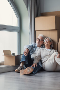 A happy senior couple relaxes on the floor beside packed moving boxes, smiling and looking out a window as they settle into their new home during retirement and relocation.