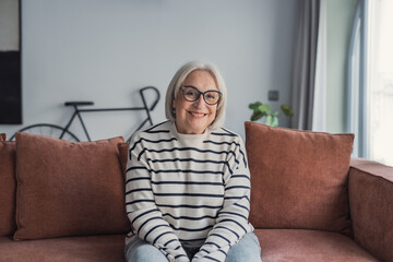 Obraz premium Smiling senior woman sits on a cozy couch in a bright living room, wearing a striped sweater and glasses, conveying warmth, comfort, confidence and a relaxed home lifestyle.