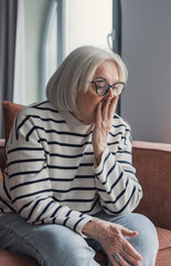 Obraz premium Elderly woman with gray hair and glasses sits on a sofa at home, appearing worried and contemplative. The image conveys loneliness, aging, concern and quiet reflection in a domestic setting.