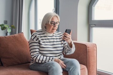 Obraz premium A senior woman sits on a comfortable couch in a bright living room, focused on her smartphone while connecting with family, managing apps and maintaining independence and everyday routines at home.