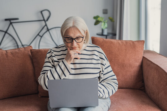 Elderly woman with glasses sits on a couch using a laptop, looking thoughtful and focused. Comfortable home setting suggests remote work, online learning, digital connectivity and independent living.