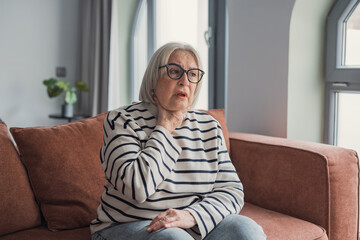 Obraz premium Elderly woman sitting on sofa holding her throat in discomfort at home