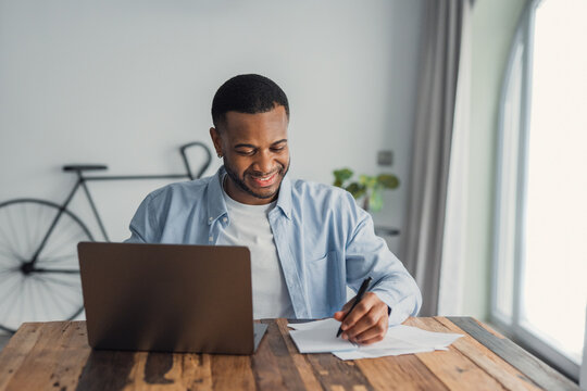 Young man smiling while working from home on a laptop and taking notes on paper at a wooden desk. Casual home office scene with natural light, productivity, focus and remote work atmosphere.