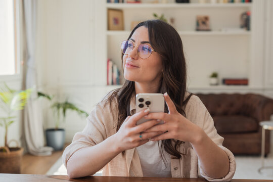 Young woman wearing glasses sits at a table in a bright living room, holding a smartphone and looking thoughtfully out the window, conveying relaxation, contemplation and casual home life.