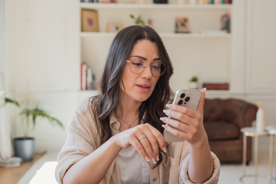 A woman in glasses uses her smartphone in a bright living room, focused on a video call or messaging. Casual remote work and everyday digital communication in a cozy home environment.