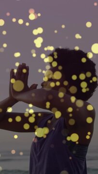 Vertical video: Woman raising arms at dusk on shore, bokeh floating over her, centering for health