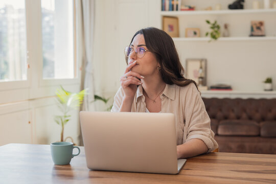 Young woman looks thoughtful while working on a laptop in a bright, cozy home office; relaxed remote work atmosphere with natural light, coffee mug, and comfortable living room background.