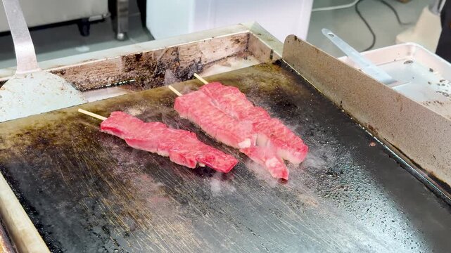 Fresh wagyu beef skewers sizzling on hot metal griddle in Japanese street food market.
