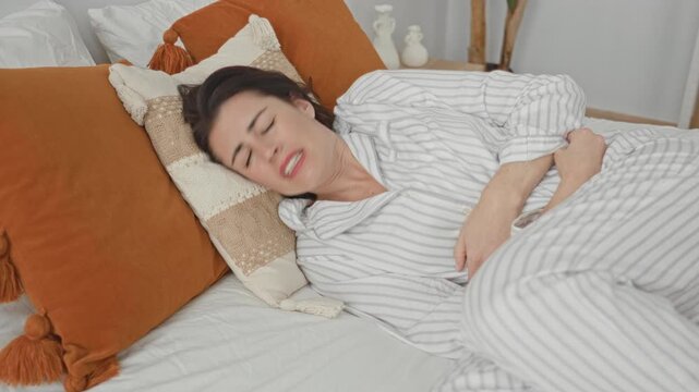 Woman clutching abdomen and holding belly while lying on a bedroom bed in striped pajamas amid orange decorative pillows and cushions; pain recovery.