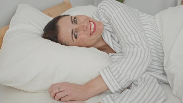 Woman lying on bed hugging a pillow and smiling with outstretched arm in building, wearing striped pajamas on white bedding; contentment morning routine.