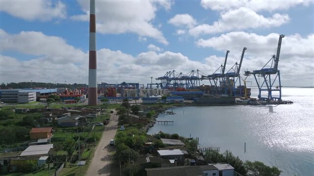 Aerial ascending shot of busy container port terminal with large cranes, stacked cargo containers, and industrial chimney beside calm waterway