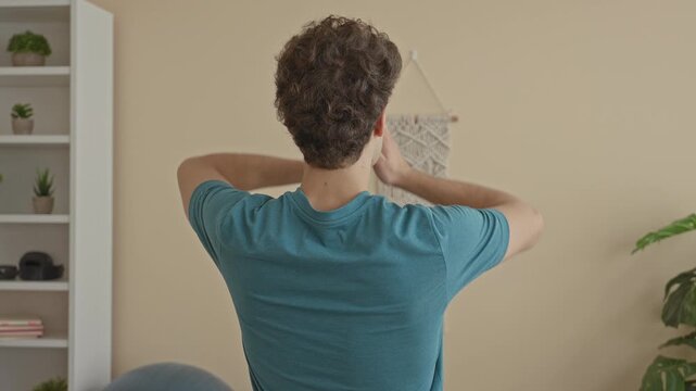Man rear view with palms together overhead practicing yoga in a studio, teal shirt and visible bare forearms, macrame wall hanging and shelf with potted plants visible; serenity.