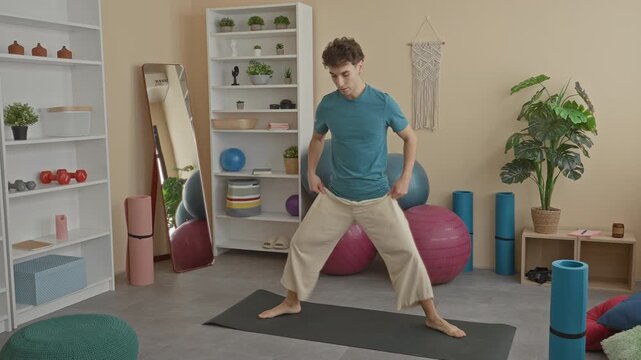 Man in teal shirt standing in wide yoga stance with arms extended and bare feet on a yoga mat in a studio; calm mindfulness.