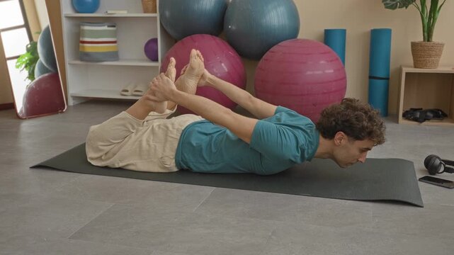 Man holds bare feet in bow pose on yoga mat surrounded by exercise balls and foam rollers in studio; serenity mindful practice.