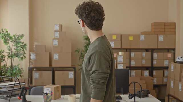 Man with packages turning head and smiling while holding a parcel and standing by stacked boxes in a building; small business optimism.