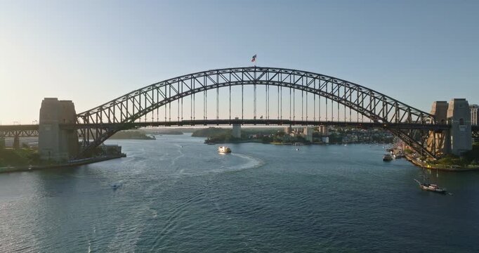 Sydney Harbour Bridge arching calm waters of the harbour, with ferry navigating the waterways and urban cityline visible in the background under a clear sky. Aerial view drone flight zoom out panorama