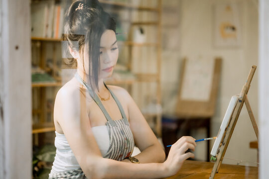 Young Asian female artist painting on canvas in cozy art studio