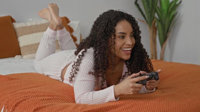 Woman lying on bed in pajamas holding a game controller with toes up, smiling while pressing buttons in building; casual joy.