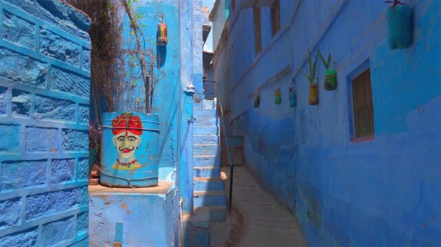 A static shot of a narrow alleyway in Jodhpur's Blue City, featuring bright blue walls, stone stairs, and decorative recycled planters with traditional Indian folk art.