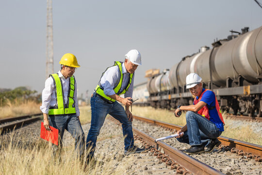 Group of asian railway engineers inspecting train tracks with freight train background, Team of professional inspectors and technicians discussing maintenance plan near oil tanker train