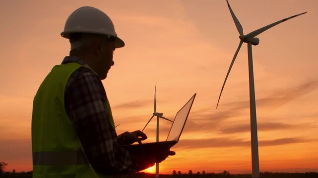 Confident male engineer in safety helmet and reflective jacket stands outdoors in a wind farm, holding a laptop and looking thoughtfully into the distance at a large wind turbine.Technician monitoring