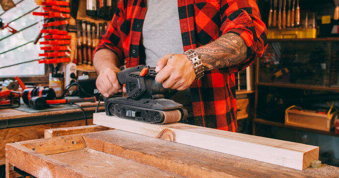 Sanding wood. Carpentry tool. Joiner man smoothing plank surface with professional belt sander equipment in garage workshop interior.