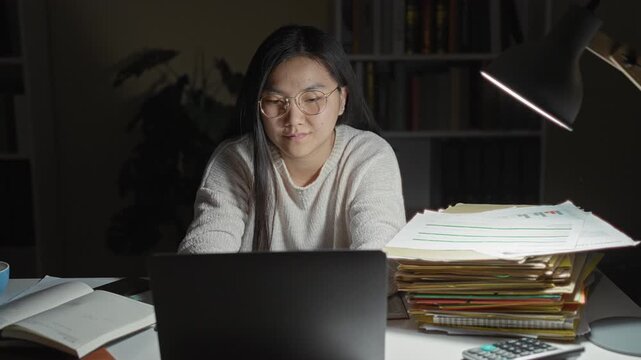 Woman at laptop with stack of folders removes glasses while rubbing nose in a building; late night study tired.
