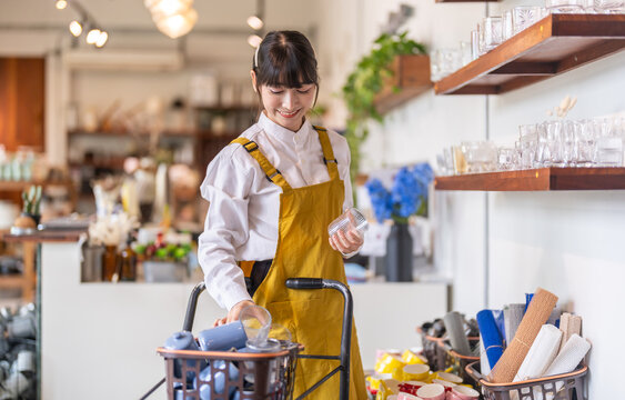 Smiling asian female small business owner in yellow apron sorting glassware in basket on utility cart, SME entrepreneur restocking shelves in home goods retail store, Lifestyle boutique management