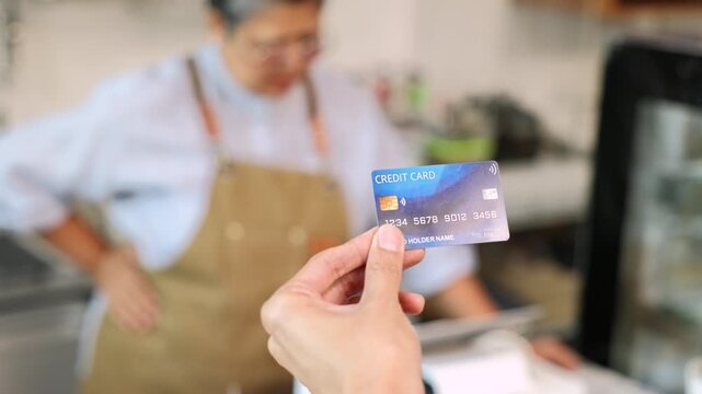 Senior entrepreneur cafe owner barista hold POS machine to customer, Asian female in apron welcome warmly in counter, croissants and glassware background, retired elderly woman start small business