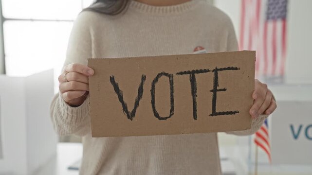Woman holds cardboard sign reading vote at a polling building with an american flag and voting booth visible; civic duty hopeful.