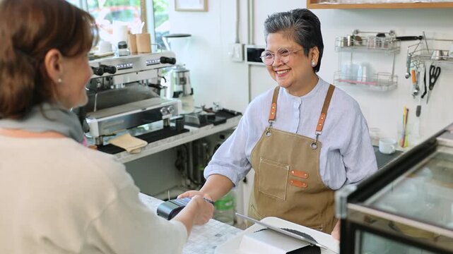 Senior entrepreneur cafe owner barista processes credit card payment on terminal, Asian female in apron smiling toward client across tiled counter, secure cashless sales inside modern coffee shop