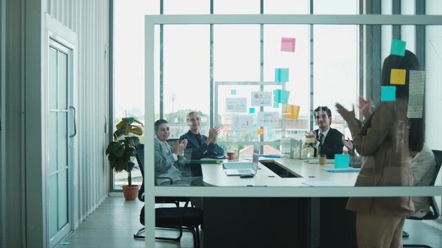 Young female company manager explains marketing investment planning strategy, by writing on glass board, presents, discusses, and brainstorms with admiration from business partner CEO team in meeting.