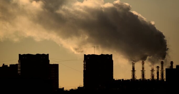 Massive steam cloud rising from multiple industrial chimneys behind dark residential buildings in warm golden backlight Dramatic urban skyline with haze district heating energy infrastructure and emis
