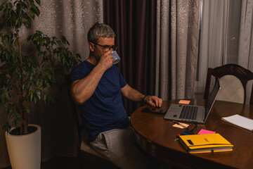Fototapeta premium Tired freelancer drinking coffee while working late at night on laptop at home