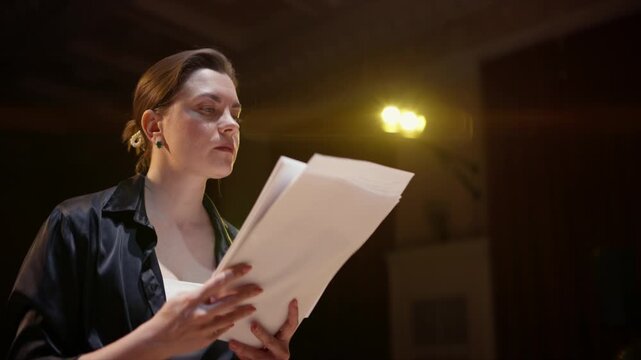 Focused woman rehearsing a dramatic script alone on a dimly lit stage, studying pages under warm theatrical lights while preparing for an emotional performance