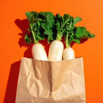Fresh white radishes with green leaves in a brown paper bag on a bright orange background