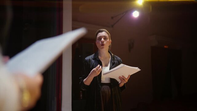 Theater director rehearsing with script pages under stage lights, guiding performers and refining dramatic performance during an evening production run-through