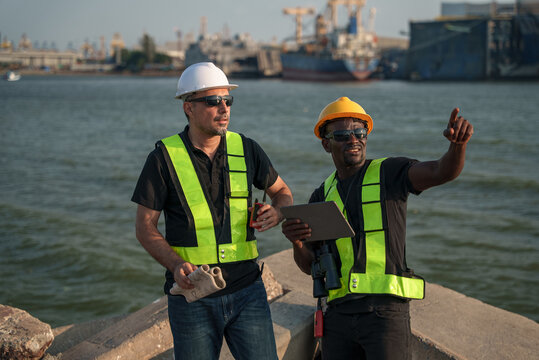 Professional marine engineer uses a radio while his colleague points toward a cargo ship at a port. Both wear safety vests and hardhats, managing logistics and industrial harbor operations