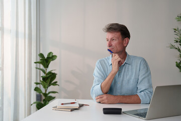 Fototapeta premium Mature man working from home, thinking and planning creative business ideas, concentrating on future projects while holding a pen at a modern white desk