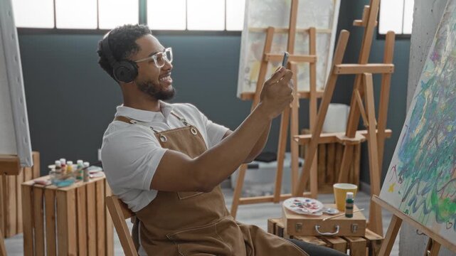 Man painter in studio sits smiling, wearing apron and headphones, holds smartphone and palette, points finger at canvas on easel; creative joy.