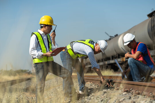 A team of railway engineers is inspecting train tracks with blueprints and radios, professional technicians in safety gear performing maintenance on rail lines.
