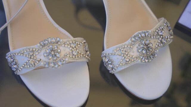 An extreme close-up showcasing the intricate rhinestone and crystal detailing on elegant white wedding shoes, resting on a reflective surface with soft-focus floral accents.