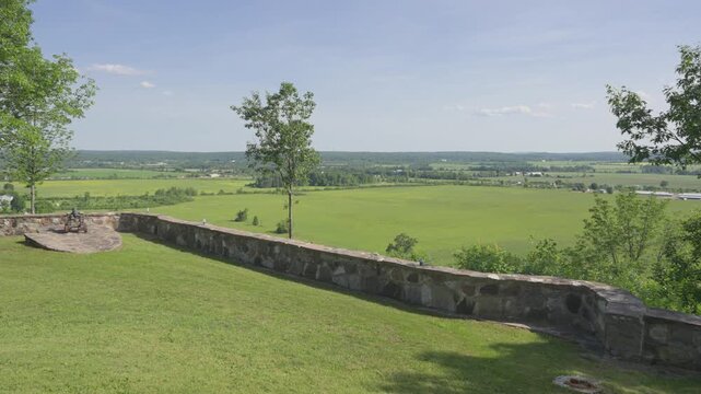 A steady wide shot of a vintage cannon on a stone lookout, featuring a long fieldstone wall and a breathtaking panoramic view of the Ottawa River Valley&rsquo;s lush farmlands.