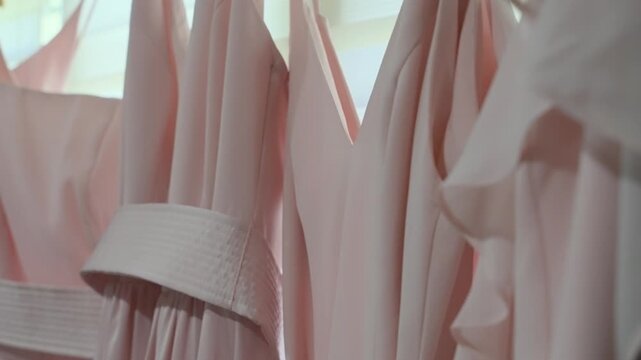 A smooth vertical tilt shot showcasing a row of matching blush pink bridesmaid dresses with various necklines and fabric details, hanging in preparation for a wedding ceremony.