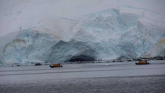 A static shot of two inflatable Zodiac boats carrying tourists in yellow parkas as they navigate the base of a massive, cracked glacier near Gamma Island, Melchior Islands