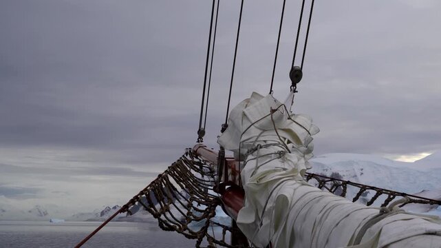 A static shot from the bow of a traditional sailing vessel as it navigates the icy waters of Antarctica's Danco Coast, featuring a wooden bowsprit and snow-covered mountains in the distance