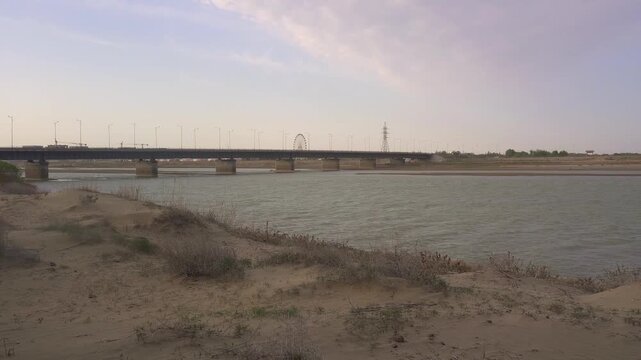 Sandy riverbank of the Syr Darya with gentle waves and a large highway bridge in the background, Kyzylorda, Kazakhstan.